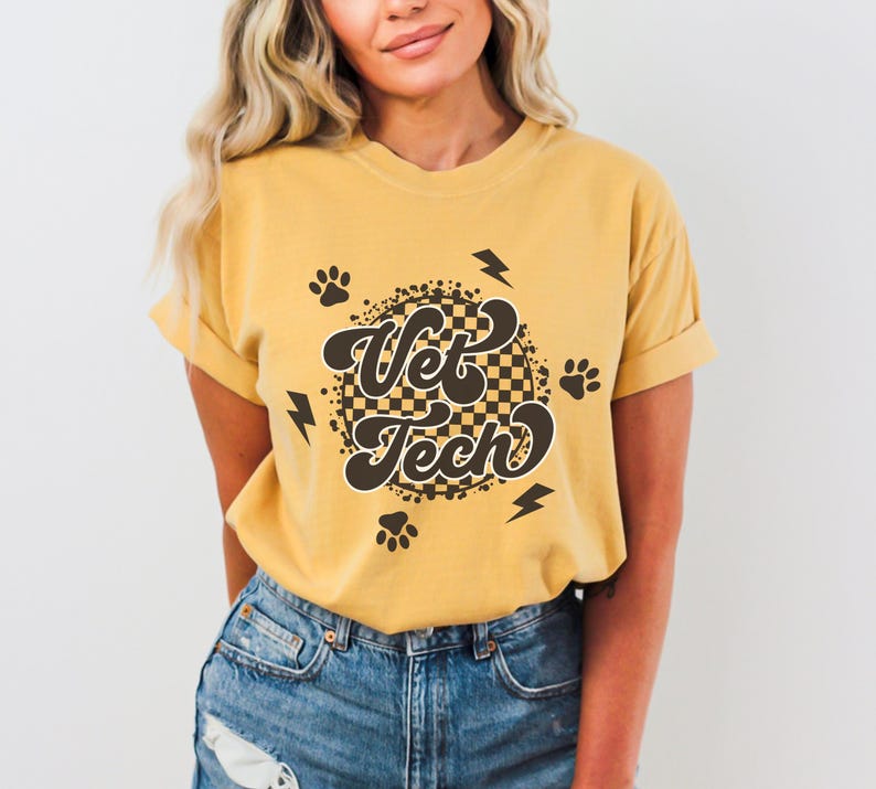 Puede incluir: Una camiseta amarilla mostaza con un gr&aacute;fico de estilo retro que dice "Vet Tech" en una fuente marr&oacute;n. El gr&aacute;fico est&aacute; rodeado de huellas de patas y rayos.