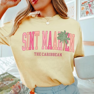 May include: A light yellow sweatshirt with the text "SINT MAARTEN" in pink block letters, with a green palm tree graphic. Below the text is "THE CARIBBEAN" in pink. The sweatshirt is a casual style.