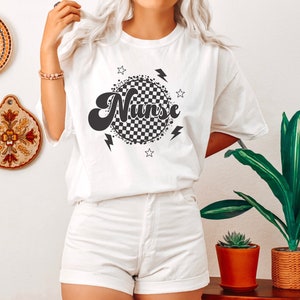 K&ouml;nnte beinhalten: Wei&szlig;es T-Shirt mit einem schwarz-wei&szlig; karierten Kreisdesign und dem Wort "Nurse" in einer Retro-Schrift. Das Design enth&auml;lt auch Sterne und Blitze.