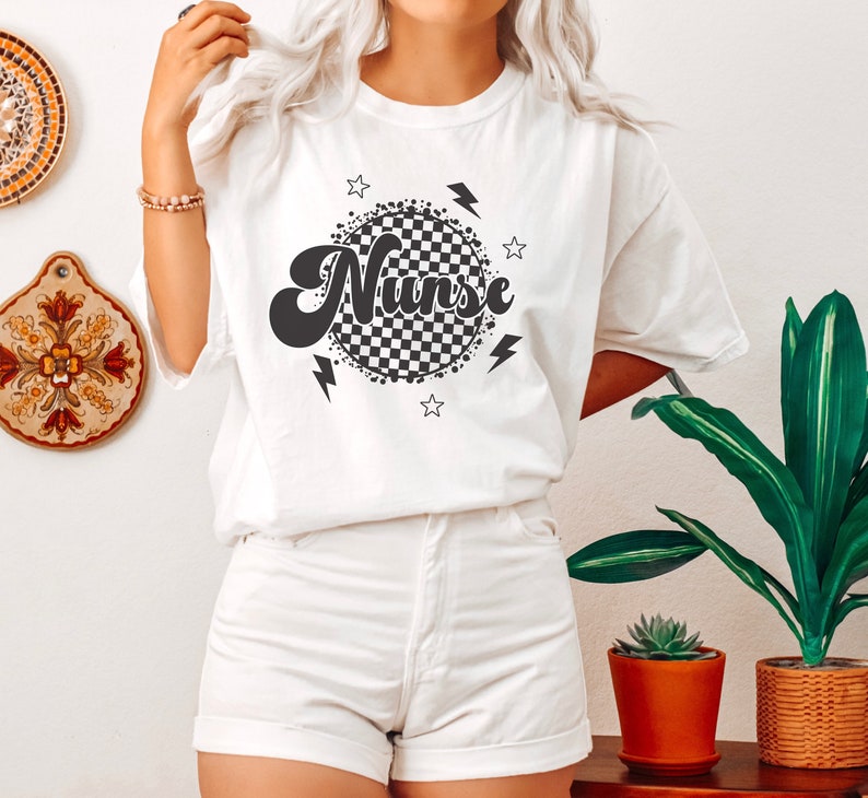 K&ouml;nnte beinhalten: Wei&szlig;es T-Shirt mit einem schwarz-wei&szlig; karierten Kreisdesign und dem Wort "Nurse" in einer Retro-Schrift. Das Design enth&auml;lt auch Sterne und Blitze.