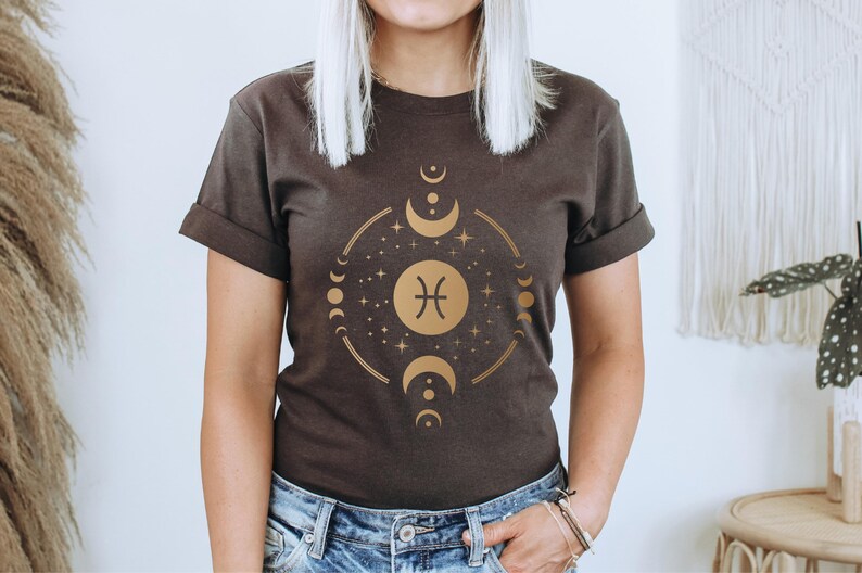 May include: A dark brown t-shirt with a gold graphic of the Pisces zodiac sign surrounded by moons and stars.