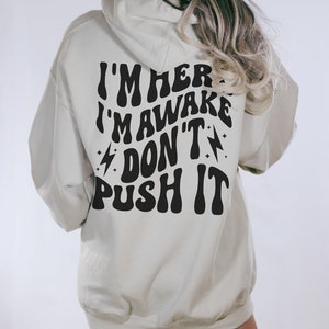May include: A light beige hoodie with a black graphic print that reads "I'm here I'm awake Don't push it" in a retro font.