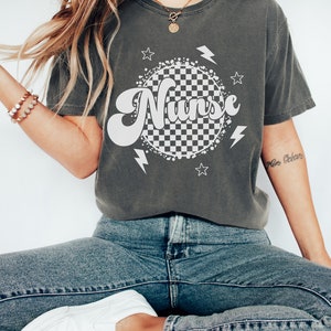 K&ouml;nnte beinhalten: Ein dunkelgraues T-Shirt mit einem wei&szlig;en karierten Kreis und dem Wort "Nurse" in einer Retro-Schrift. Der Kreis ist von wei&szlig;en Sternen und Blitzen umgeben.