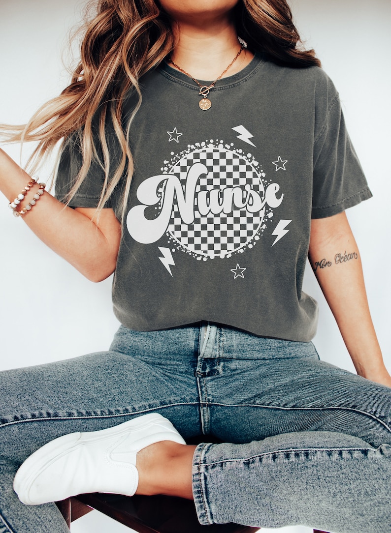 K&ouml;nnte beinhalten: Ein dunkelgraues T-Shirt mit einem wei&szlig;en karierten Kreis und dem Wort "Nurse" in einer Retro-Schrift. Der Kreis ist von wei&szlig;en Sternen und Blitzen umgeben.