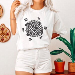 Puede incluir: Camiseta blanca con un dise&ntilde;o circular de cuadros blancos y negros que dice "Vet Tech" en una fuente retro. El dise&ntilde;o tambi&eacute;n incluye huellas de patas y rayos.