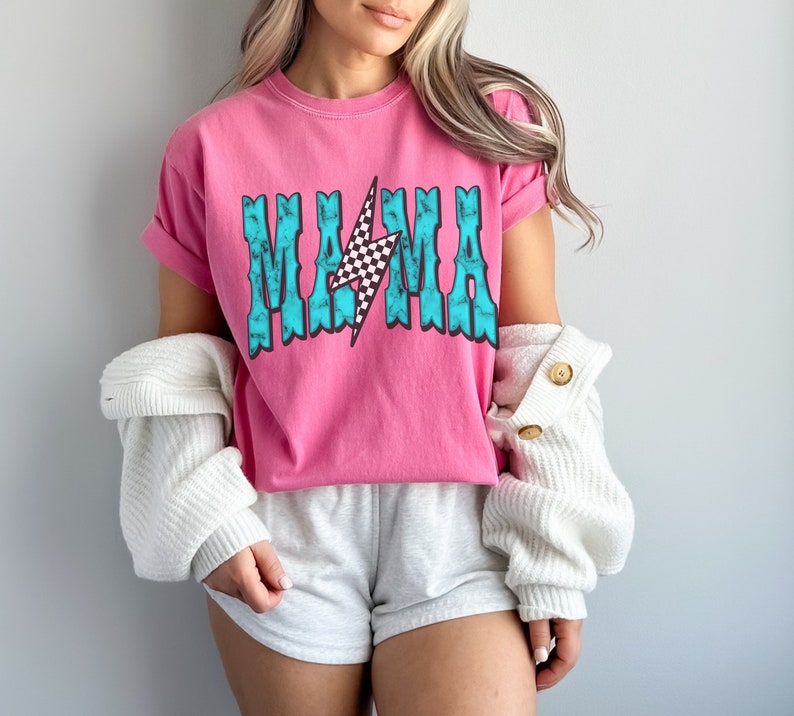 Puede incluir: Camiseta rosa de manga corta con un gr&aacute;fico azul turquesa que dice "MAMA" con un rayo a cuadros en el centro.