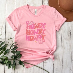 May include: Pink t-shirt with a retro design featuring the text "Howdy Howdy Howdy" in pink and white with a cowboy hat and boot graphic.