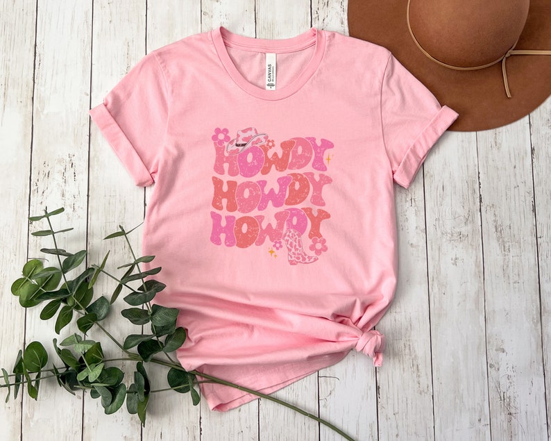 May include: Pink t-shirt with a retro design featuring the text "Howdy Howdy Howdy" in pink and white with a cowboy hat and boot graphic.