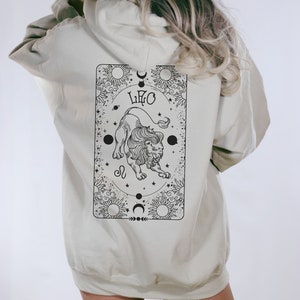May include: A beige hoodie with a black and white graphic on the back. The graphic features a lion, the word "LEO", and celestial elements. The hoodie has a hood and long sleeves.