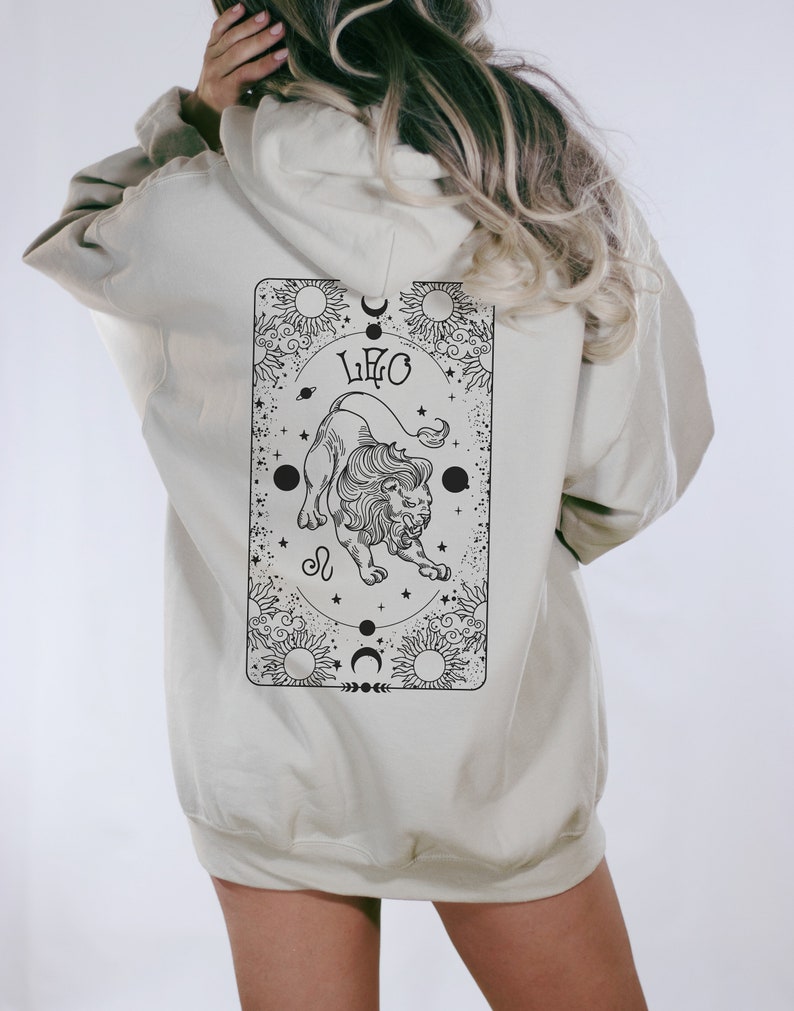 May include: A beige hoodie with a black and white graphic on the back. The graphic features a lion, the word "LEO", and celestial elements. The hoodie has a hood and long sleeves.