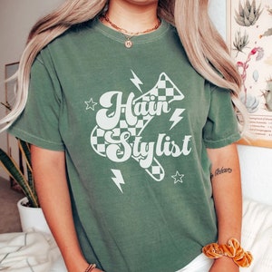 May include: A green t-shirt with a white graphic of a checkered megaphone with the text "Hair Stylist" in a retro font.