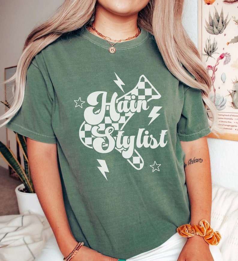 May include: A green t-shirt with a white graphic of a checkered megaphone with the text "Hair Stylist" in a retro font.