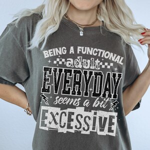 May include: Dark grey t-shirt with the text "BEING A FUNCTIONAL adult EVERYDAY seems a bit EXCESSIVE" in black and white block letters. The shirt has short sleeves and a crew neck.