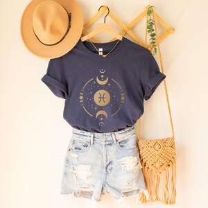 May include: A navy blue t-shirt with a gold zodiac symbol and moon phases graphic. The shirt is styled with denim shorts and a straw bag.