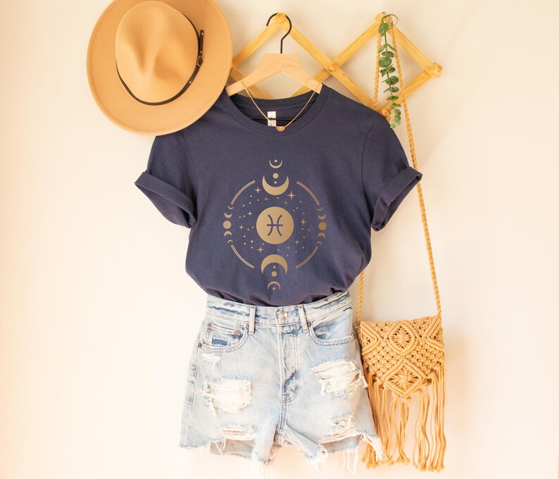 May include: A navy blue t-shirt with a gold zodiac symbol and moon phases graphic. The shirt is styled with denim shorts and a straw bag.