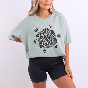 Puede incluir: Una camiseta verde claro con un dise&ntilde;o circular de cuadros negros y blancos que dice "Vet Tech" en una fuente retro. El dise&ntilde;o tambi&eacute;n incluye huellas de patas y rayos.