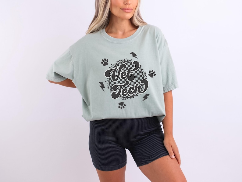 Puede incluir: Una camiseta verde claro con un dise&ntilde;o circular de cuadros negros y blancos que dice "Vet Tech" en una fuente retro. El dise&ntilde;o tambi&eacute;n incluye huellas de patas y rayos.