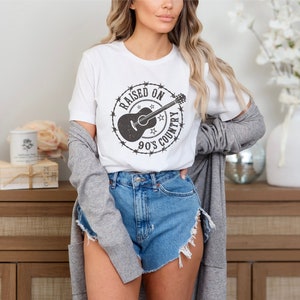 May include: White t-shirt with a distressed black graphic featuring a guitar and the text "RAISED ON 90'S COUNTRY" inside a barbed wire circle. The shirt is paired with denim shorts and a gray cardigan.