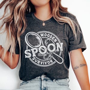 May include: A dark gray t-shirt with a white graphic of a wooden spoon in a circle with the text "Wooden Spoon Survivor".