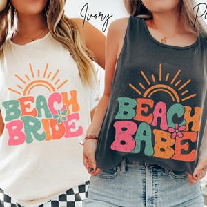May include: Two sleeveless shirts, one ivory and one dark gray, each with a colorful graphic. The ivory shirt says "Beach Bride" with a sun and flower design. The dark gray shirt says "Beach Babe" with a sun and flower design.