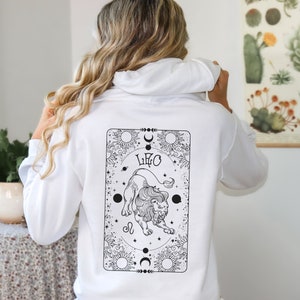 May include: White hoodie with a black and white graphic of a lion and the word "LEO" on the back. The design is framed by celestial elements like suns, moons, and stars. The hoodie is a casual garment.