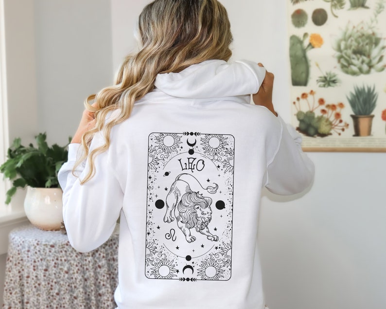 May include: White hoodie with a black and white graphic of a lion and the word "LEO" on the back. The design is framed by celestial elements like suns, moons, and stars. The hoodie is a casual garment.
