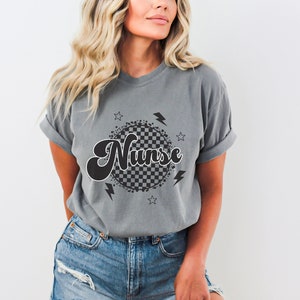 K&ouml;nnte beinhalten: Graues T-Shirt mit einem schwarz-wei&szlig;en karierten Kreisdesign und dem Wort "Nurse" in einer Retro-Schrift. Das Design enth&auml;lt auch Sterne und Blitze.