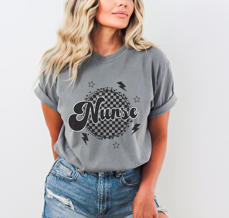 K&ouml;nnte beinhalten: Graues T-Shirt mit einem schwarz-wei&szlig;en karierten Kreisdesign und dem Wort "Nurse" in einer Retro-Schrift. Das Design enth&auml;lt auch Sterne und Blitze.
