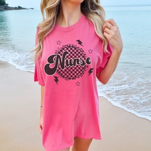 K&ouml;nnte beinhalten: Rosa, &uuml;bergro&szlig;es T-Shirt mit einem schwarzen und wei&szlig;en karierten Kreis-Motiv und dem Wort "Nurse" in einer Retro-Schrift.