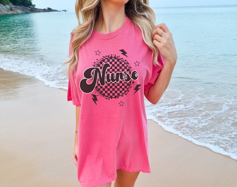K&ouml;nnte beinhalten: Rosa, &uuml;bergro&szlig;es T-Shirt mit einem schwarzen und wei&szlig;en karierten Kreis-Motiv und dem Wort "Nurse" in einer Retro-Schrift.