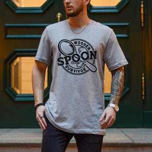 May include: A gray t-shirt with a black and white graphic design of a wooden spoon. The graphic design includes the text "Wooden Spoon Survivor".