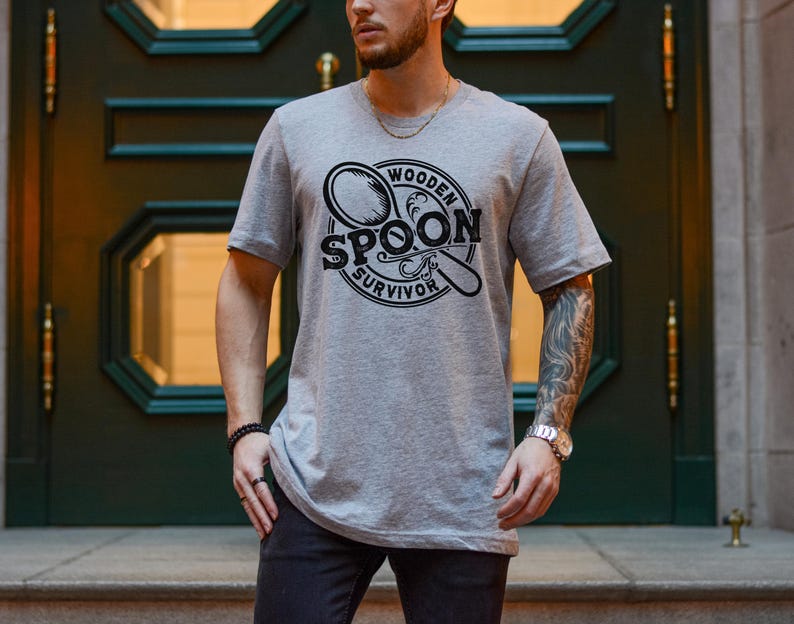 May include: A gray t-shirt with a black and white graphic design of a wooden spoon. The graphic design includes the text "Wooden Spoon Survivor".