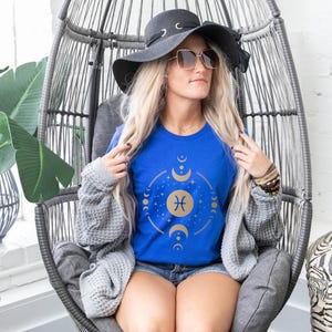 May include: A woman wearing a blue t-shirt with a gold Pisces zodiac symbol and moon phases graphic. She is wearing a gray cardigan and a wide-brimmed black hat with a gold chain.