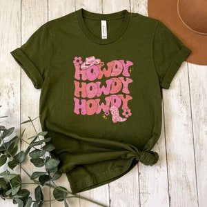 May include: A green t-shirt featuring a pink and white graphic that says "Howdy" three times. The graphic also includes a pink cowboy hat and boots.
