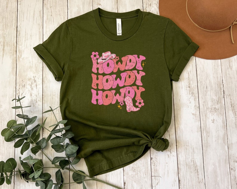 May include: A green t-shirt featuring a pink and white graphic that says "Howdy" three times. The graphic also includes a pink cowboy hat and boots.