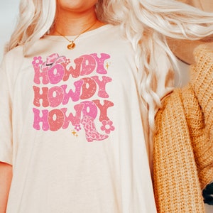 May include: A white t-shirt featuring a pink graphic design that reads "Howdy" three times. The graphic also includes a pink cowboy hat and a pink cowboy boot with a floral design.