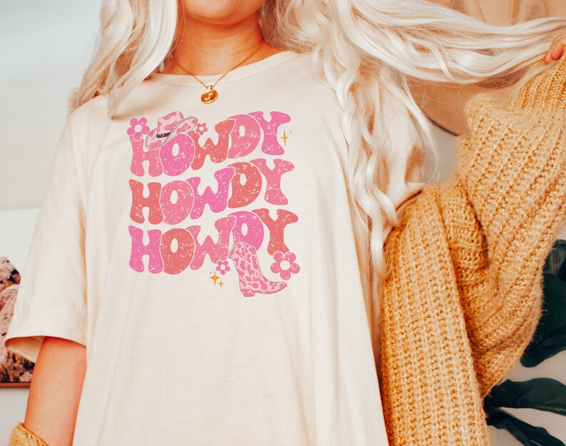 May include: A white t-shirt featuring a pink graphic design that reads "Howdy" three times. The graphic also includes a pink cowboy hat and a pink cowboy boot with a floral design.