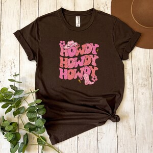 May include: A brown t-shirt featuring a pink and white graphic that reads "Howdy" three times. The graphic includes a cowboy hat and boot.