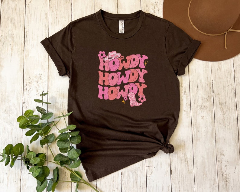 May include: A brown t-shirt featuring a pink and white graphic that reads "Howdy" three times. The graphic includes a cowboy hat and boot.