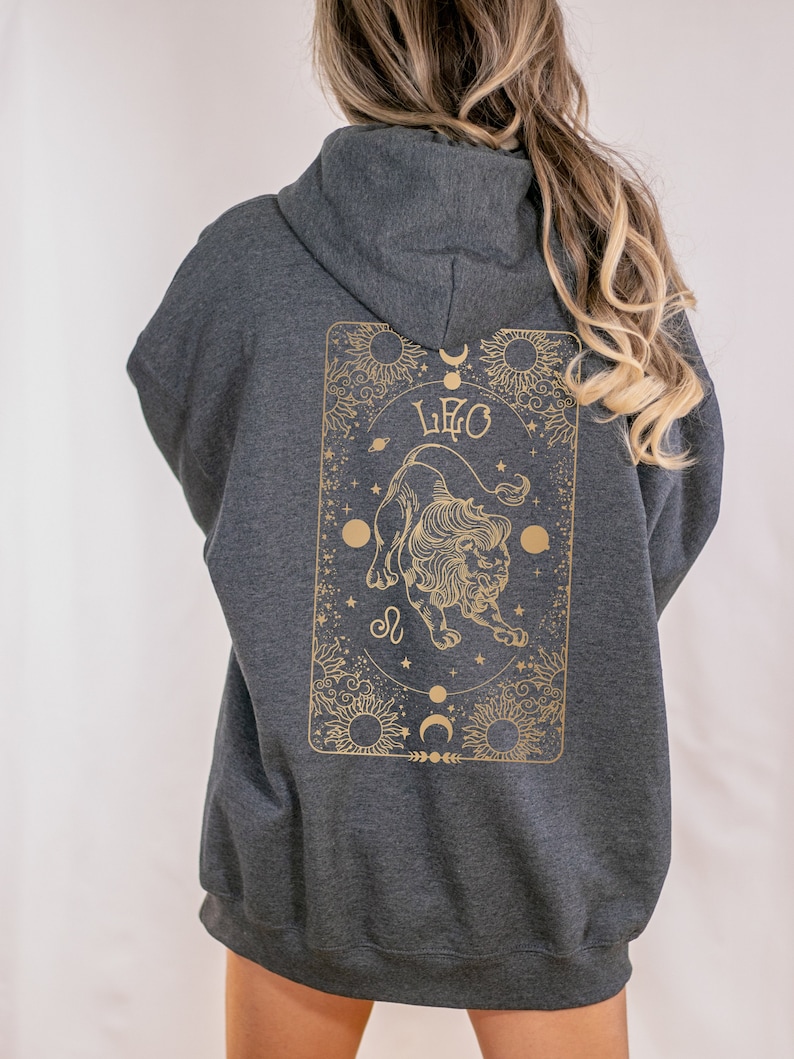 May include: Dark gray hoodie with a gold Leo zodiac design on the back. The design features a lion, suns, moons, and stars. The hoodie has a hood and long sleeves.