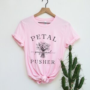May include: Pink t-shirt with a black graphic that says "PETAL professional PUSHER" and a bouquet of flowers.