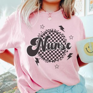 K&ouml;nnte beinhalten: Pinkes T-Shirt mit einem schwarz-wei&szlig;en karierten Kreisdesign und dem Wort "Nurse" in einer Retro-Schrift.