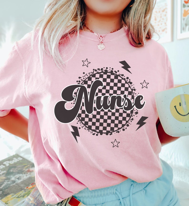K&ouml;nnte beinhalten: Pinkes T-Shirt mit einem schwarz-wei&szlig;en karierten Kreisdesign und dem Wort "Nurse" in einer Retro-Schrift.