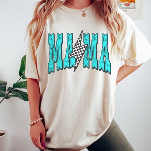 Puede incluir: Una camiseta beige claro con un gr&aacute;fico azul turquesa que dice "MAMA" con un rayo a cuadros en el medio.