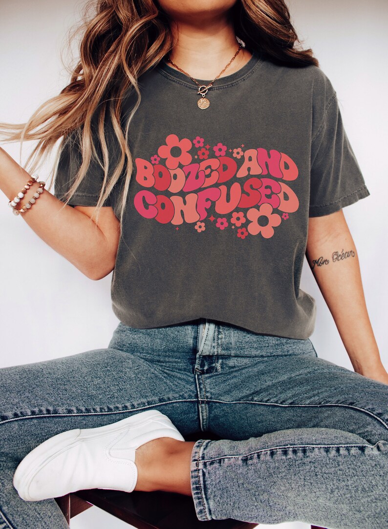 May include: A dark grey t-shirt with a retro-style graphic that says "Boozed and Confused" in pink and red letters with flowers surrounding the text.