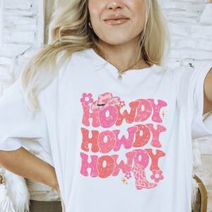 May include: White t-shirt featuring a pink and red graphic design that says "Howdy" three times. The graphic also includes a pink cowboy hat and a pink and brown cowboy boot.
