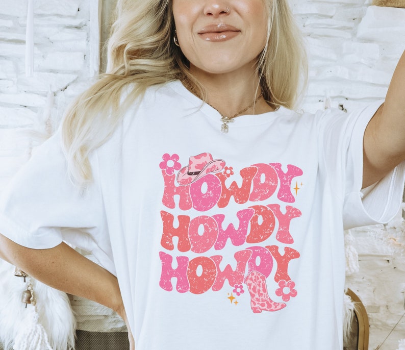 May include: White t-shirt featuring a pink and red graphic design that says "Howdy" three times. The graphic also includes a pink cowboy hat and a pink and brown cowboy boot.