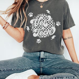 Puede incluir: Una camiseta gris oscuro con un dise&ntilde;o gr&aacute;fico blanco con las palabras "Vet Tech" en una fuente retro. El gr&aacute;fico tambi&eacute;n incluye huellas de patas y rayos.