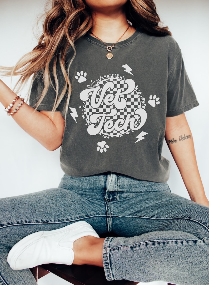 Puede incluir: Una camiseta gris oscuro con un dise&ntilde;o gr&aacute;fico blanco con las palabras "Vet Tech" en una fuente retro. El gr&aacute;fico tambi&eacute;n incluye huellas de patas y rayos.