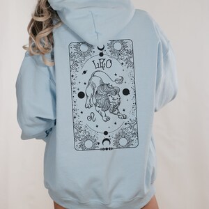 May include: Light blue hoodie with a black Leo zodiac design on the back. The design features a lion, the word "LEO", suns, moons, and stars. The hoodie has a hood and long sleeves.